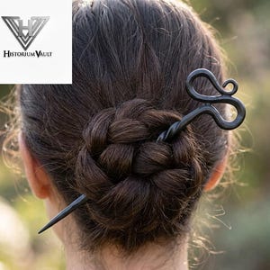 May include: A dark metal hair pin with a decorative swirl design is inserted into a braided hairstyle. The pin is a dark gray color and has a pointed end. The logo for Historium Vault is in the upper left corner.