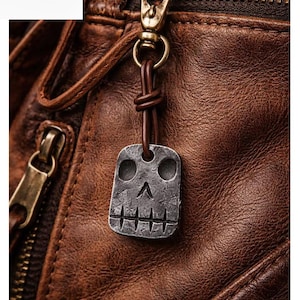 Könnte beinhalten: Nahaufnahme einer braunen Lederjacke mit einem Totenkopf-Metallanhänger, der an einem Messingreißverschluss hängt. Der Anhänger ist quadratisch mit einem Totenkopf-Design. Die Jacke hat sichtbare Nähte und ein Logo oben links.