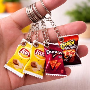 Op de afbeelding: Miniatuur sleutelhanger set met vier snackzakjes: twee gele Lays zakjes, een rode Doritos zak en een oranje Cheetos zak. De zakjes zijn bevestigd aan een zilveren sleutelhangerring. De Lays zakjes tonen chips, terwijl de Doritos zak nacho cheese chips toont.