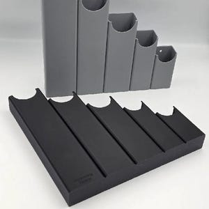 Puede incluir: Un conjunto de cinco tubos de almacenamiento verticales grises con parte superior abierta y una base rectangular horizontal negra con cinco ranuras paralelas de longitudes variables. La base negra tiene el texto "Avancing Design" en relieve.