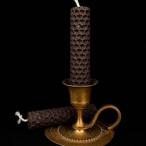 Könnte beinhalten: Eine dunkelbraune Kerze mit Wabenmuster und weißem Docht steht in einem Vintage-Kerzenhalter aus Messing. Eine weitere Kerze mit dem gleichen Design liegt daneben. Der Hintergrund ist schwarz.