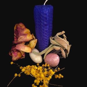 Op de afbeelding: Een cilindervormige blauwe kaars met honingraatstructuur en een witte lont, vergezeld door gedroogde gele mimosa-bloemen, gedroogde rozenblaadjes, een gladde witte steen, een ronde roze steen en gedroogde saliebladeren.