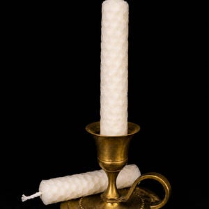 Peut inclure: Un bougeoir en laiton doré supporte une haute bougie crème à motif en nid d'abeille. Une deuxième bougie de même conception repose à côté du bougeoir. Le fond est noir, mettant en valeur les tons chauds de la bougie et du support.