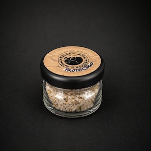 Peut inclure: Petit pot en verre rempli de sel rituel. Le couvercle noir porte l'inscription "PROTECTION" en blanc, avec "SEL RITUEL RITUAL SALT" dans un motif circulaire. Le sel est un mélange de blanc et de vert. Le pot est sur une surface noire.