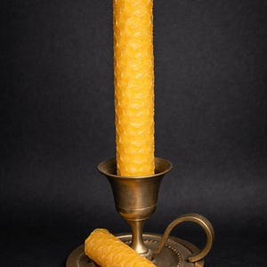 Puede incluir: Una vela alta de cera de abeja amarilla con un patrón de panal de abeja se encuentra en un candelabro de latón antiguo. Una vela más pequeña y similar está en la base. Fondo negro.