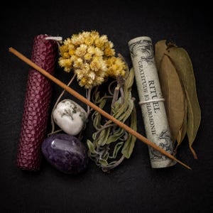 Peut inclure: Composition spirituelle sur fond noir. Comprend une bougie bordeaux, des fleurs jaunes séchées, une pierre blanche et grise, une pierre violette, de la sauge, un rouleau de papier avec du texte, un bâton d'encens brun et des feuilles séchées.