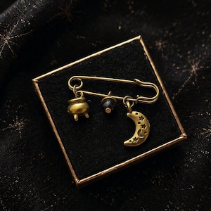 Peut inclure: Épingle de sûreté dorée avec trois breloques : un chaudron, une perle sombre et un croissant de lune étoilé. L'épingle est présentée dans un coffret noir avec un bord doré, sur un tissu noir à motifs d'étoiles dorées.