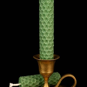 Può includere: Una candela verde a nido d'ape è in un candelabro vintage in ottone, con una seconda candela accanto. Le candele hanno una superficie strutturata e uno stoppino bianco. Lo sfondo è nero.