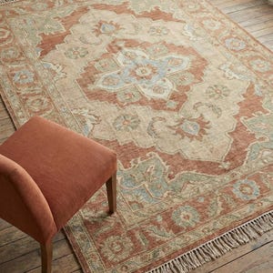 Könnte beinhalten: Rechteckiger Teppich mit floralem Muster in Beige-, Rost- und Blautönen. Der Teppich hat einen Fransenrand und liegt auf einem Holzboden. Ein rostfarbener Stuhl befindet sich im Vordergrund.
