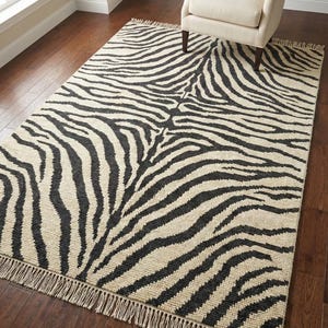 Puede incluir: Una alfombra rectangular con un estampado de rayas de cebra en negro y beige. La alfombra tiene un borde con flecos y está colocada sobre un suelo de madera oscura. Un sillón beige está en el fondo.