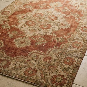 May include: A rectangular rug with a distressed appearance and a floral design. The rug has a central area of faded red with beige floral patterns and a beige border with red floral details. It is placed on a light-coloured tiled floor.