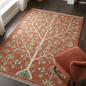 Puede incluir: Alfombra de color terracota con un diseño de árbol crema con detalles florales azules y verdes. La alfombra tiene un borde con flecos y está colocada sobre un suelo de madera oscura. Una silla de color óxido está en primer plano.