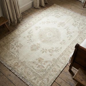 Pode incluir: Tapete retangular creme com um padrão floral e borda com franjas. O tapete apresenta um design de medalhão central e motivos florais em tons de bege e castanho. O tapete está colocado sobre um chão de madeira.