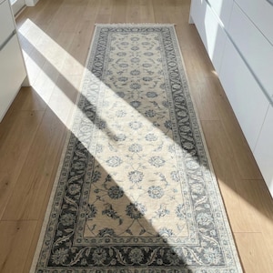 May include: A rectangular beige runner rug with a floral pattern and a dark gray border. The rug has a fringe on both ends and is placed on a light wood floor. The rug's dimensions are approximately 3 feet wide and 8 feet long.