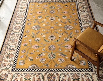 May include: A rectangular area rug featuring a mustard yellow centre with a symmetrical floral vine pattern in pink, green, and white. The rug has a wide cream border with intricate floral motifs, a black and white striped trim, and white fringe.