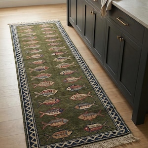 Puede incluir: Una alfombra verde con un diseño de peces. La alfombra tiene un borde azul oscuro y crema con flecos. Los peces son de varios colores, incluyendo rojo, azul y marrón. La alfombra está colocada sobre un suelo de madera.