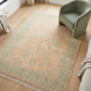May include: A rectangular area rug with a peach-colored center and a sage green border. The rug features a floral pattern and a fringed edge. The rug is placed on a hardwood floor, with a green armchair visible in the background.