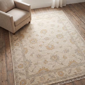 Op de afbeelding: Een rechthoekig beige vloerkleed met een bloemenpatroon in goud en grijs. Het vloerkleed heeft een rand en franjes langs de randen. Een beige fauteuil staat in de linkerbovenhoek en het vloerkleed ligt op een houten vloer.