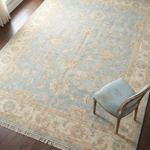 Puede incluir: Una alfombra rectangular grande con un campo central azul claro y un borde beige. La alfombra presenta un intrincado diseño floral en tonos beige y naranja. Una silla azul claro está sobre la alfombra.