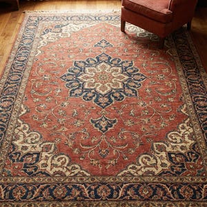 Puede incluir: Una alfombra rectangular y ornamentada con un diseño central de medallón. La alfombra presenta un campo rojo terracota con un borde y acentos azul marino y crema. La alfombra tiene un borde con flecos y está colocada sobre un suelo de madera.