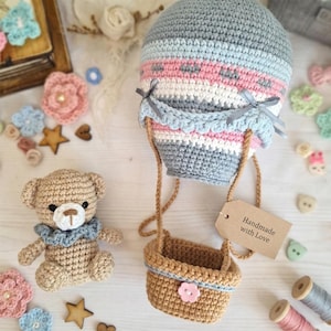 May include: Crocheted hot air balloon in gray, pink, and blue with a brown basket, and a small crocheted teddy bear. The balloon has a tag that reads "Handmade with Love". Decorative flowers and buttons surround the objects.