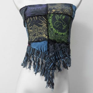 Peut inclure: Haut tube patchwork avec divers motifs célestes et géométriques dans les tons de bleu, vert et or. Le haut a une frange en bas. Le haut est présenté sur un mannequin blanc.