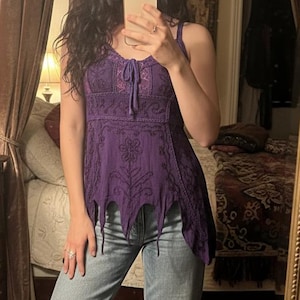 Może przedstawiać: Fioletowy top na ramiączkach z misternym haftem i unikalnym, asymetrycznym dołem. Połączony z jasnoniebieskimi dżinsami. Obraz uchwycony w lustrze, odbijający pokój z wystrojem w stylu vintage.