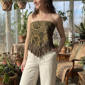 May include: A strapless top with a sun and celestial design in gold and olive green, paired with cream-colored linen pants. The top has a fringed hem. The setting is a sunroom with hanging plants and vintage furniture.