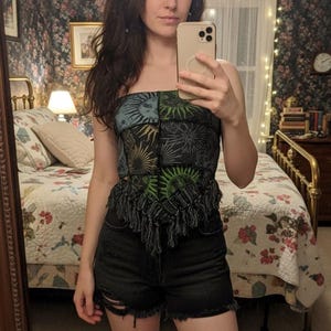 May include: A strapless top with a sun and moon pattern in blue, green, and black, paired with black denim shorts. The top has a fringed hemline. The image is taken in a bedroom with a floral wallpaper and a mirror.
