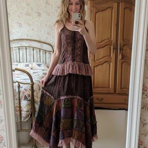 May include: A brown patchwork maxi dress with spaghetti straps, a lace-trimmed neckline, and a tiered skirt. The dress features embroidered details and a patchwork design in various colours. The model is holding a phone and standing in front of a mirror.