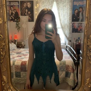 Può includere: Un top senza maniche verde acqua con dettagli ricamati e un orlo smerlato unico. Il top presenta un dettaglio con lacci sullo scollo. L'immagine è riflessa in uno specchio dorato decorativo e ornato.