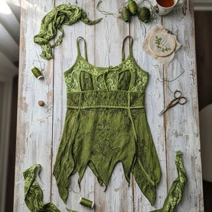 Può includere: Un top verde senza maniche con un design floreale e dettagli in pizzo, appoggiato su una superficie di legno rustica imbiancata. Il top è accompagnato da ritagli di tessuto verde, rocchetti di filo, un telaio da ricamo, forbici e una tazza di tè.