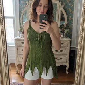 Könnte beinhalten: Olivgrünes Tanktop mit aufwendigen gestickten Blumenmustern und einem Schnürhalsausschnitt. Das Oberteil hat einen einzigartigen, wellenförmigen Saum und wird mit weißen Shorts kombiniert. Das Bild wird in einem Spiegel aufgenommen.