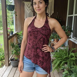 Könnte beinhalten: Burgunderfarbenes Neckholder-Top mit Sonnen- und Blumenmuster. Das Oberteil hat einen asymmetrischen Saum und wird mit Jeansshorts kombiniert. Die Person trägt ein Armband mit mehreren Perlen.