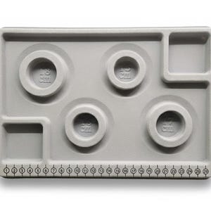 Peut inclure: Un plateau gris clair pour bijoux avec plusieurs compartiments et une règle le long du bord inférieur. Le plateau comporte quatre renfoncements circulaires étiquetés "18 CM" et un petit compartiment carré.