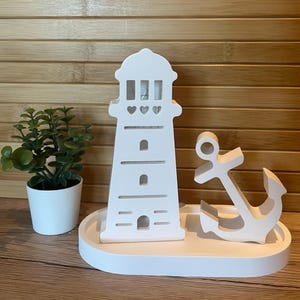 May include: A white nautical-themed decorative set featuring a lighthouse, an anchor, and a small potted plant. The lighthouse has heart-shaped cutouts and rectangular windows. The set is displayed on a white oval tray.