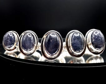 May include: Five silver rings with oval-shaped blue gemstones. The rings are arranged in a row on a black surface.