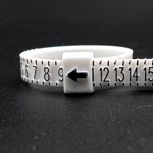 May include: White plastic ring sizer with black numbers 6 through 15 and an arrow pointing to the left.
