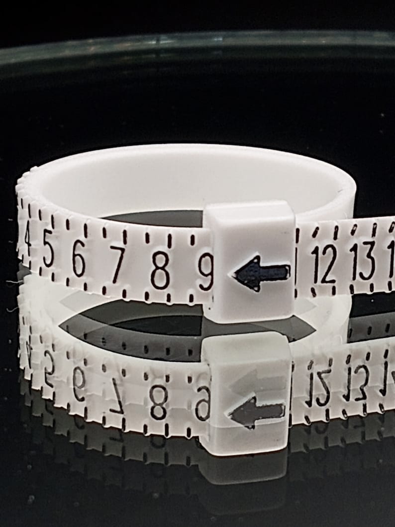 May include: White plastic ring sizer with black markings and arrows. The numbers on the sizer range from 4 to 15.