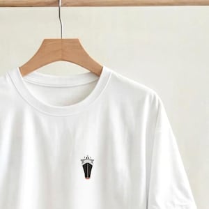 May include: White crew neck t-shirt with a small graphic of a ship on the front. The ship illustration is black, white, and red. The t-shirt is hanging on a wooden hanger.