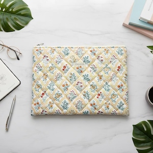 May include: A quilted laptop sleeve with a floral pattern in yellow, orange, and blue. The sleeve is rectangular with a zipper closure. The image also includes a notebook, pen, glasses, and a cup of coffee.