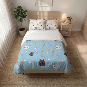Puede incluir: Un edredón azul claro y dos fundas de almohada a juego con un patrón repetitivo de caras de gato en blanco y negro, lunas crecientes amarillas, corazones rosas, estrellas coloridas y varitas. El juego de cama se muestra sobre una cama de madera.