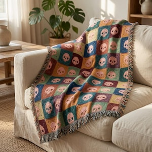 Puede incluir: Una manta tejida con un patrón de cuadrícula repetitivo, donde cada cuadrado contiene un motivo de calavera estilizada. La manta presenta una paleta multicolor de tonos azul verdoso, amarillo mostaza, burdeos y rosa, con bordes deshilachados.