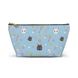Puede incluir: Un estuche con cremallera de color azul claro que presenta un patrón repetitivo de caras de gato blancas, caras de gato negras, lunas crecientes amarillas, corazones rosas, estrellas multicolores y varitas rosas con detalles dorados. Cremallera dorada.