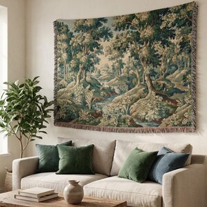 Puede incluir: Un tapiz que representa una serena escena de bosque con un río, árboles y follaje en tonos verdes, beige y azul. El tapiz está adornado con flecos y cuelga sobre un sofá beige con cojines verdes y azules. Una mesa de centro de madera y una planta en maceta están en primer plano.