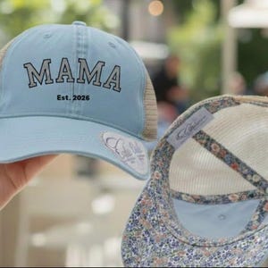 Puede incluir: Gorra de camionero azul claro con la palabra "MAMA" en letras negras y "Est. 2026". La gorra tiene una parte trasera de malla y un interior con estampado floral. Se ve una segunda gorra, que muestra el estampado floral.