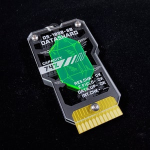 Könnte beinhalten: Ein transparenter Datenspeicher aus Kunststoff mit einem grünen, juwelenförmigen Symbol. Der Speicher hat einen goldenen Stecker und den Text "05-1898-68 DATASHARD" und "KAPAZITÄT 74%". Der Text "RES.CHK-OK E.FIELD-OK DATA.UP-OK INT.CHK-....." ist ebenfalls sichtbar.