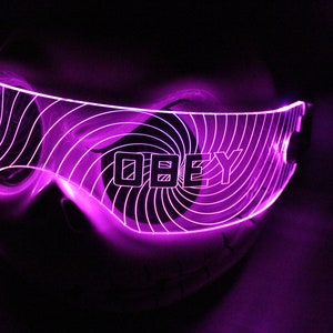 Peut inclure: Lunettes LED violettes avec un motif en spirale et le mot "OBEY" au centre. Les lunettes sont éclairées, créant un effet lumineux sur un fond sombre. Le design est futuriste et accrocheur.