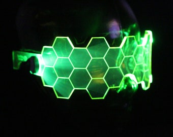 Peut inclure: Lunettes lumineuses vertes à LED avec un motif en nid d'abeille. Les lunettes sont portées par une personne dans une pièce sombre.