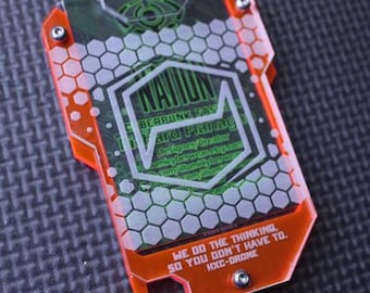 May include: A bright orange and green plastic badge with a hexagonal pattern. The badge has a black and white logo with the text "NATION", "BERPUNK FAS", "Fard Flamونغ", "Designer Greator", "WE DO THE THINKING, SO YOU DON'T HAVE TO.", and "HXC-DRONE".